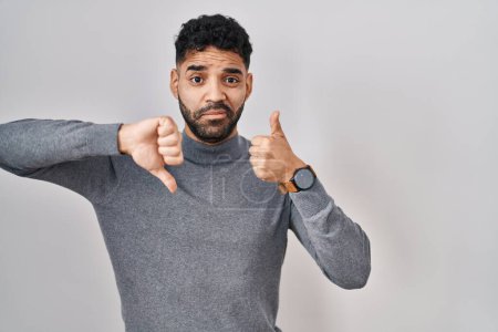 Foto de Hispanic man with beard standing over white background doing thumbs up and down, disagreement and agreement expression. crazy conflict - Imagen libre de derechos