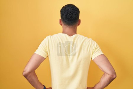 Foto de Hispanic man with beard standing over yellow background standing backwards looking away with arms on body - Imagen libre de derechos