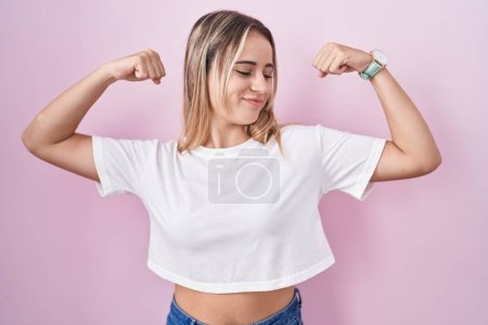 Téléchargez les photos : Jeune femme blonde debout sur fond rose montrant les muscles des bras souriant fier. concept de fitness. - en image libre de droit