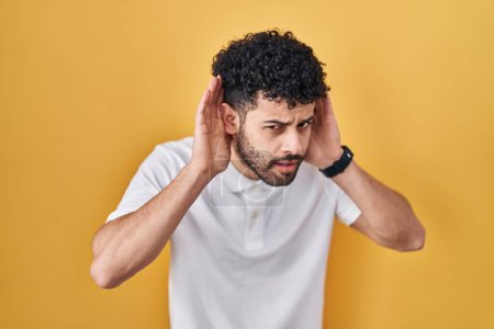Photo for Arab man standing over yellow background trying to hear both hands on ear gesture, curious for gossip. hearing problem, deaf - Royalty Free Image