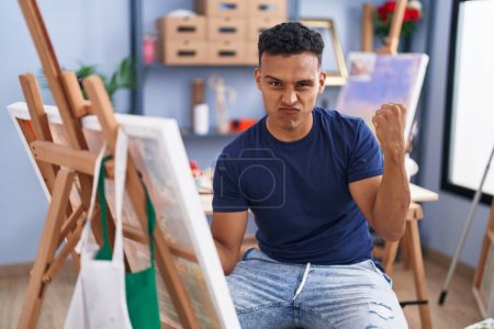 Photo for Young hispanic man painting sitting at art studio screaming proud, celebrating victory and success very excited with raised arms - Royalty Free Image