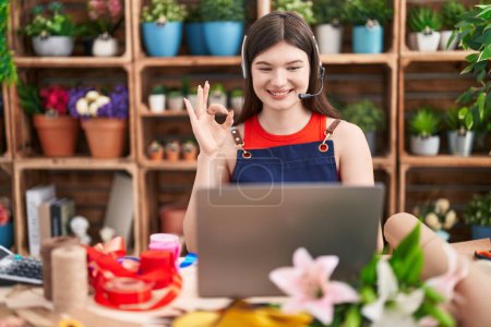 Téléchargez les photos : Jeune femme caucasienne travaillant au magasin de fleuriste faire appel vidéo faire ok signe avec les doigts, sourire geste amical excellent symbole - en image libre de droit