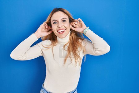 Téléchargez les photos : Jeune femme caucasienne debout sur fond bleu souriant tirant les oreilles avec les doigts, geste drôle. problème d'audition - en image libre de droit