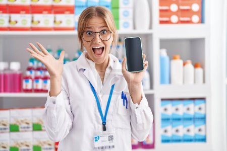 Photo for Young caucasian woman working at pharmacy drugstore showing smartphone screen celebrating victory with happy smile and winner expression with raised hands - Royalty Free Image