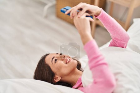 Foto de Joven mujer hispana hermosa usando teléfono inteligente acostado en la cama en el dormitorio - Imagen libre de derechos