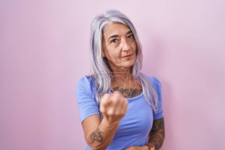 Foto de Mujer de mediana edad con tatuajes de pie sobre fondo rosa haciendo señas ven aquí gesto con la mano invitando a la bienvenida feliz y sonriente - Imagen libre de derechos