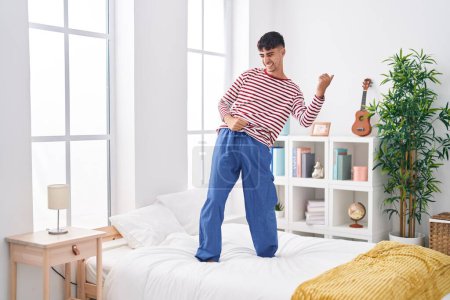 Téléchargez les photos : Jeune homme hispanique souriant danse confiante sur le lit dans la chambre - en image libre de droit