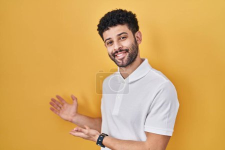 Foto de Hombre árabe de pie sobre fondo amarillo invitando a entrar sonriente natural con la mano abierta - Imagen libre de derechos