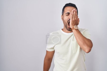 Foto de Hombre hispano con barba de pie sobre un fondo aislado bostezando cansado cubriendo media cara, ojos y boca con la mano. cara duele en el dolor. - Imagen libre de derechos