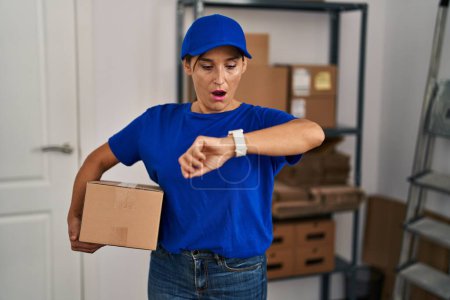 Téléchargez les photos : Femme brune du moyen âge travaillant en uniforme de livraison et chapeau regardant le temps de la montre inquiet, peur de se retrouver en retard - en image libre de droit