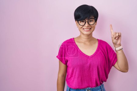 Téléchargez les photos : Jeune femme asiatique avec les cheveux courts debout sur fond rose montrant et pointant vers le haut avec le doigt numéro un tout en souriant confiant et heureux. - en image libre de droit