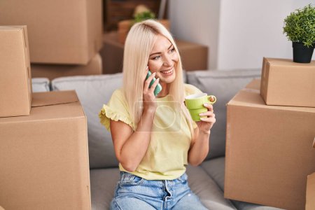 Photo for Young blonde woman talking on the smartphone drinking coffee at new home - Royalty Free Image