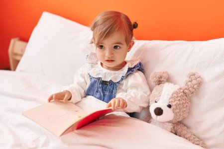 Téléchargez les photos : Adorable livre de lecture blond pour tout-petit assis sur le lit avec un ours en peluche dans la chambre - en image libre de droit