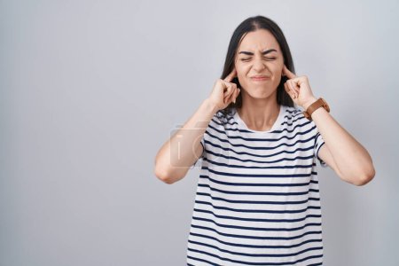 Photo for Young brunette woman wearing striped t shirt covering ears with fingers with annoyed expression for the noise of loud music. deaf concept. - Royalty Free Image