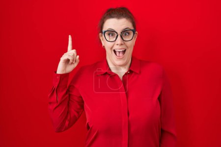 Téléchargez les photos : Jeune femme hispanique aux cheveux roux debout sur fond rouge pointant du doigt avec une idée réussie. Sorti et heureux. numéro un. - en image libre de droit
