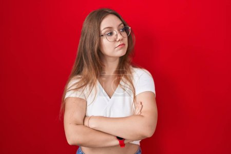 Foto de Young caucasian woman standing over red background looking to the side with arms crossed convinced and confident - Imagen libre de derechos