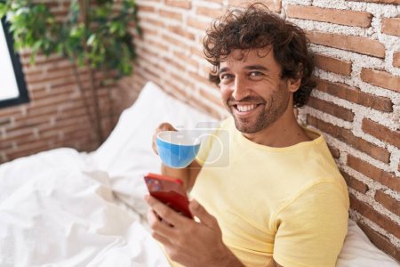 Photo for Young hispanic man using smartphone drinking coffee at bedroom - Royalty Free Image