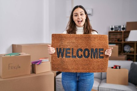 Photo for Young hispanic woman holding welcome doormat at new home celebrating crazy and amazed for success with open eyes screaming excited. - Royalty Free Image