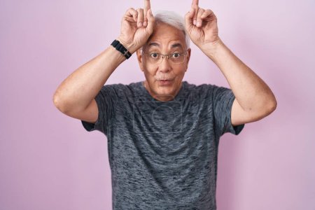 Téléchargez les photos : Homme d'âge moyen avec les cheveux gris debout sur fond rose faisant geste drôle avec doigt sur la tête comme cornes de taureau - en image libre de droit