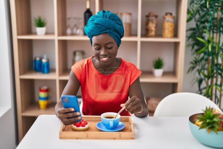 Foto de Mujer afroamericana joven usando smartphone desayunando en casa - Imagen libre de derechos
