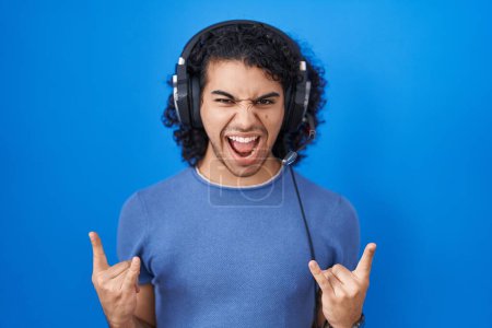 Téléchargez les photos : Hispanic man with curly hair listening to music using headphones shouting with crazy expression doing rock symbol with hands up. music star. heavy concept. - en image libre de droit