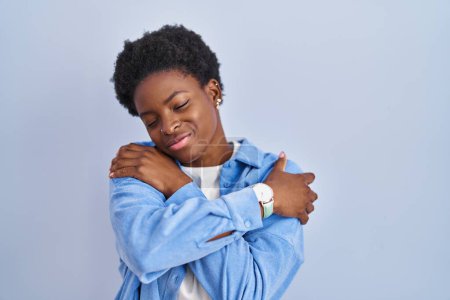 Photo for African american woman standing over blue background hugging oneself happy and positive, smiling confident. self love and self care - Royalty Free Image