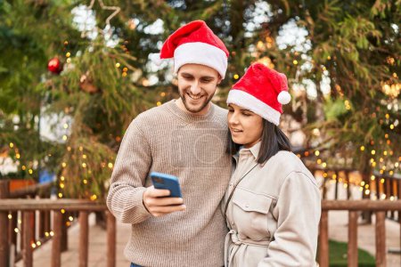 Téléchargez les photos : Couple homme et femme portant un chapeau de Noël en utilisant un smartphone au parc - en image libre de droit