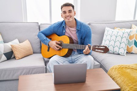 Foto de Hombre hispano joven que tiene clase de guitarra clásica en línea sentado en el sofá en casa - Imagen libre de derechos