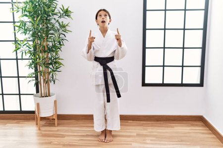 Foto de Young hispanic girl wearing karate kimono and black belt amazed and surprised looking up and pointing with fingers and raised arms. - Imagen libre de derechos
