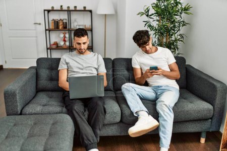 Foto de Dos hombres hispanos en pareja usando smartphone y laptop en casa - Imagen libre de derechos