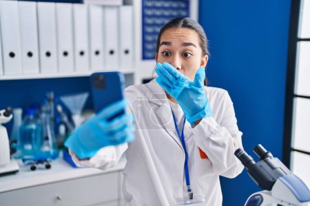 Téléchargez les photos : Young south asian woman working at scientist laboratory with smartphone covering mouth with hand, shocked and afraid for mistake. surprised expression - en image libre de droit
