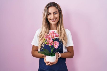 Foto de Mujer rubia joven con delantal de jardinero sosteniendo planta con una sonrisa feliz y fresca en la cara. persona afortunada. - Imagen libre de derechos