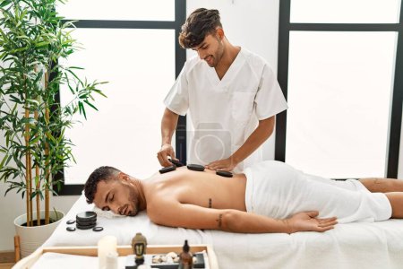Photo for Two hispanic men physiotherapist and patient having back treatment using hot stones at beauty center - Royalty Free Image