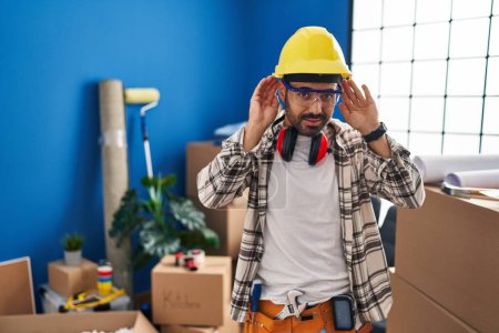 Téléchargez les photos : Jeune homme hispanique avec barbe travaillant à la rénovation de la maison en essayant d'entendre les deux mains sur le geste de l'oreille, curieux de commérages. problème auditif, sourd - en image libre de droit