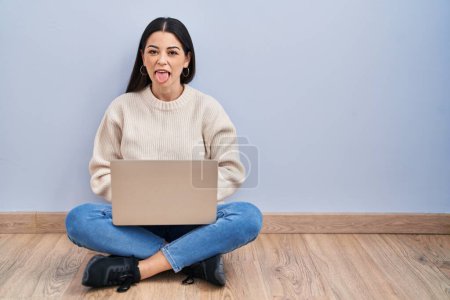 Foto de Mujer joven usando el ordenador portátil sentado en el suelo en casa sacando la lengua feliz con expresión divertida. concepto de emoción. - Imagen libre de derechos
