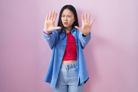 Foto de Young asian woman standing over pink background doing stop gesture with hands palms, angry and frustration expression - Imagen libre de derechos