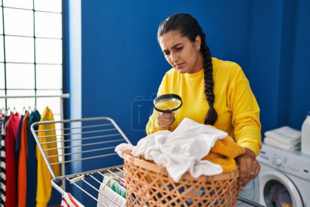 Foto de Mujer hispana joven colgando ropa en tendedero usando lupa en la lavandería - Imagen libre de derechos