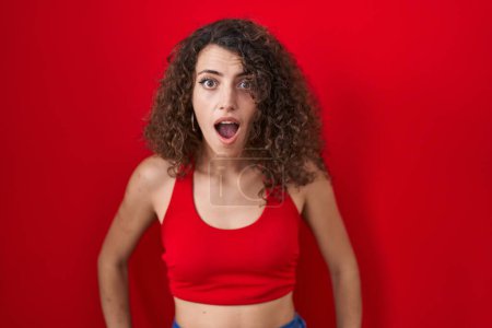 Téléchargez les photos : Hispanic woman with curly hair standing over red background afraid and shocked with surprise and amazed expression, fear and excited face. - en image libre de droit