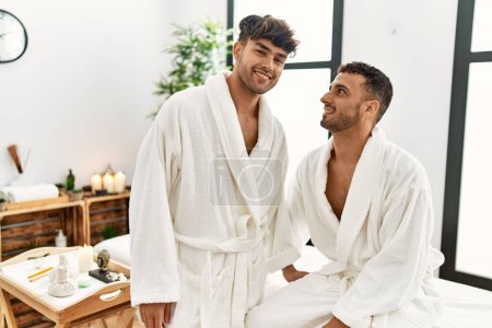 Téléchargez les photos : Two hispanic men couple wearing bathrobe sitting on massage table at beauty center - en image libre de droit