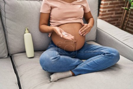 Téléchargez les photos : Jeune femme latine enceinte appliquant lotion crème sur le ventre de la peau à la maison - en image libre de droit