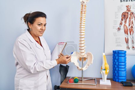 Téléchargez les photos : Femme hispanique du moyen âge physiothérapeute pointant vers le modèle anatomique de colonne vertébrale à la clinique de réadaptation - en image libre de droit