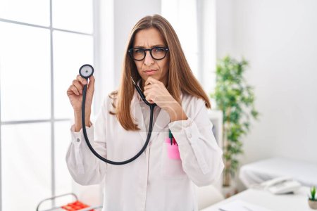 Photo for Young woman wearing doctor uniform holding stethoscope serious face thinking about question with hand on chin, thoughtful about confusing idea - Royalty Free Image