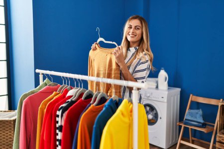 Photo for Young blonde woman smiling confident holding clothes of rack at laundry room - Royalty Free Image