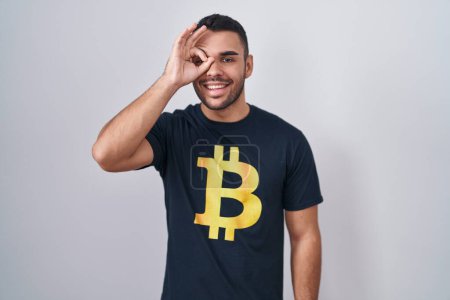 Téléchargez les photos : Jeune homme hispanique portant Bitcoin t-shirt faire un geste correct avec la main souriante, oeil regardant à travers les doigts avec un visage heureux. - en image libre de droit