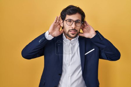 Téléchargez les photos : Beau latin homme debout sur fond jaune essayant d'entendre les deux mains sur le geste de l'oreille, curieux de commérages. problème auditif, sourd - en image libre de droit
