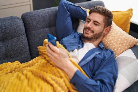 Téléchargez les photos : Jeune homme caucasien utilisant smartphone couché sur le canapé à la maison - en image libre de droit