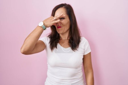 Foto de Middle age brunette woman standing over pink background smelling something stinky and disgusting, intolerable smell, holding breath with fingers on nose. bad smell - Imagen libre de derechos