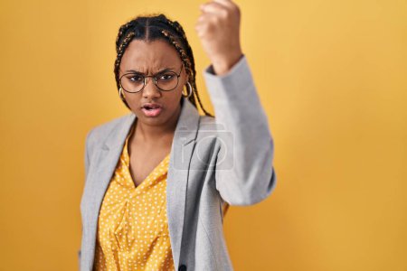 Photo for African american woman with braids standing over yellow background angry and mad raising fist frustrated and furious while shouting with anger. rage and aggressive concept. - Royalty Free Image