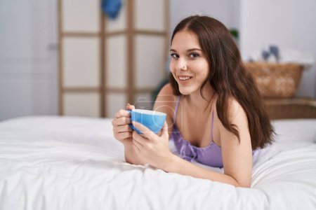 Foto de Young woman drinking cup of coffee lying on bed at bedroom - Imagen libre de derechos