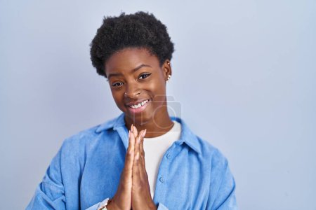 Foto de African american woman standing over blue background praying with hands together asking for forgiveness smiling confident. - Imagen libre de derechos